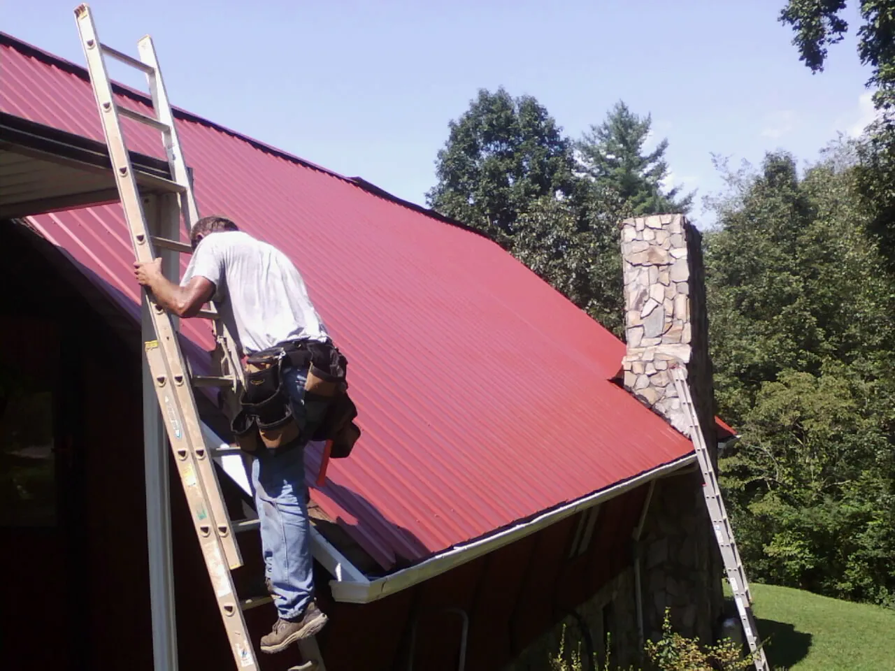 Roofer on a ladder beside a red metal roof and stone chimney — Soffit & Fascia Repair by Roofing Brothers in Delaware